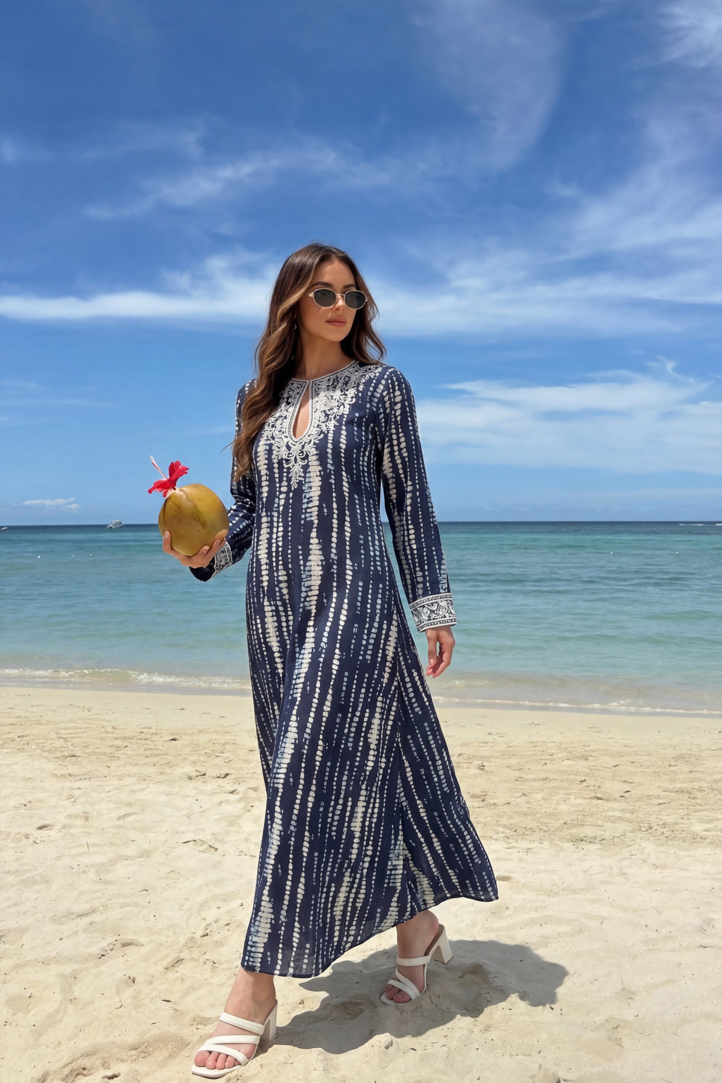 Woman in a long dress holding a coconut on a beach with blue sky and ocean.