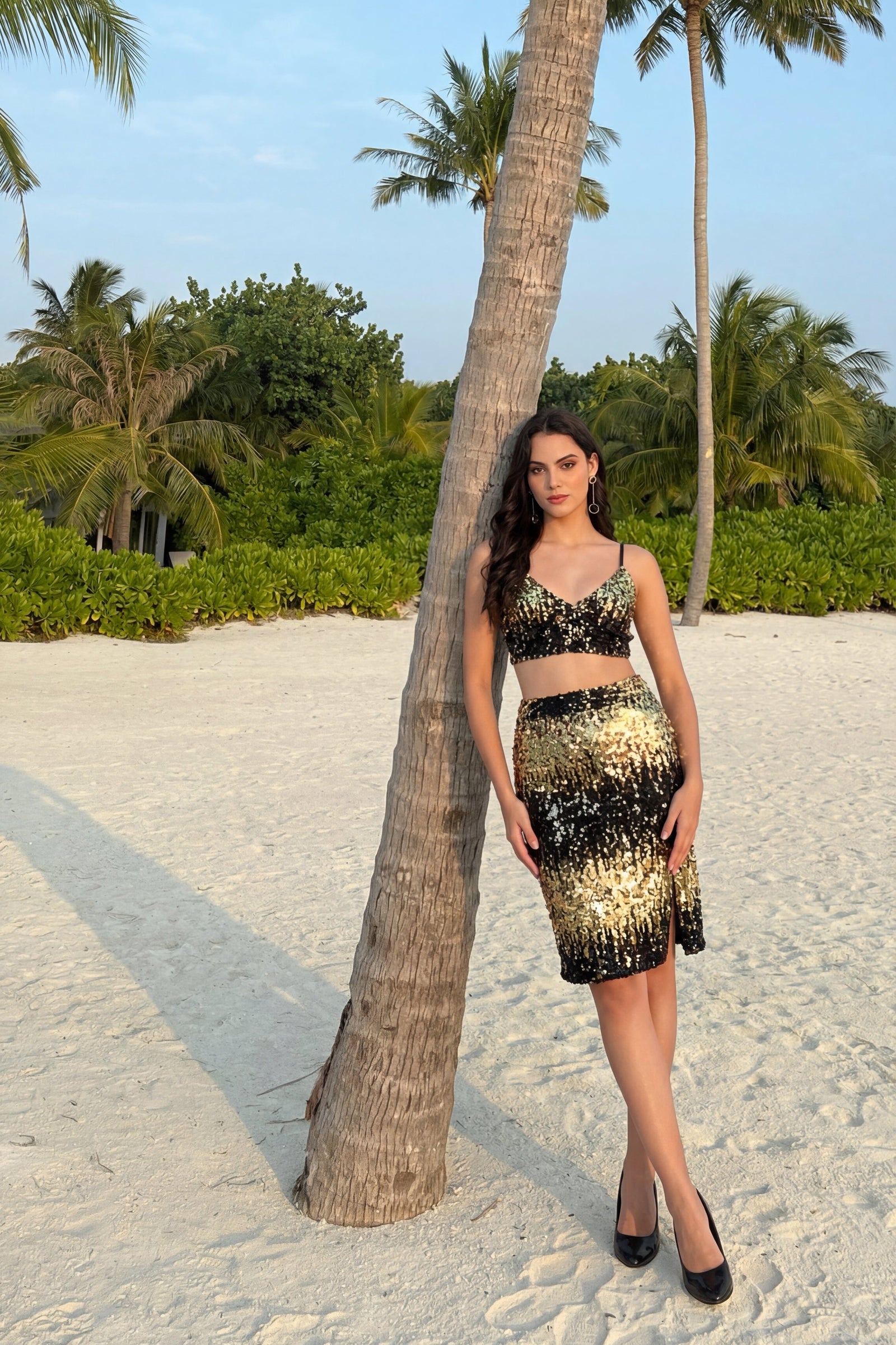 Woman in a black and gold dress standing next to a palm tree on a sandy beach.