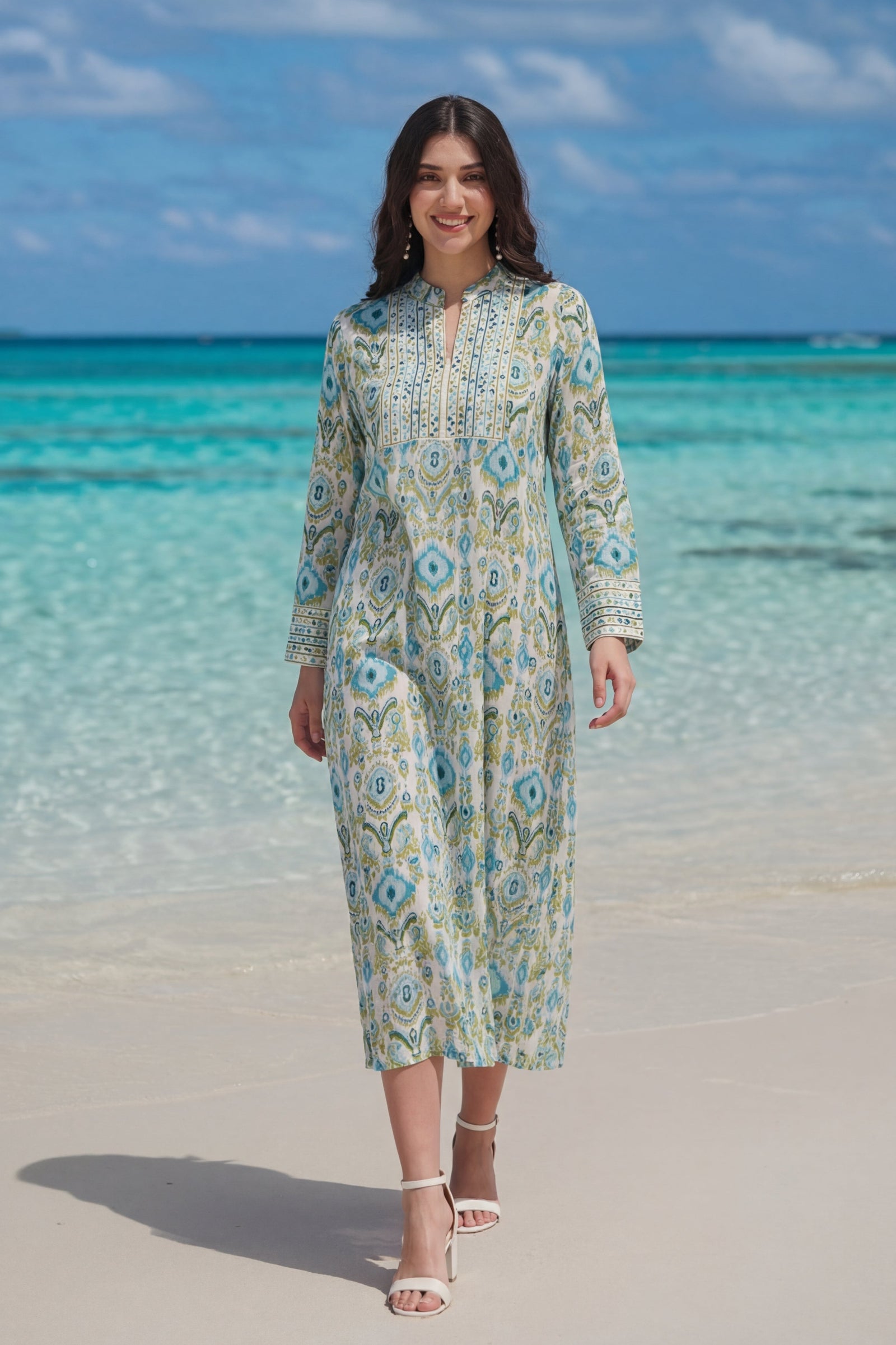 Woman in a floral dress standing on a beach with clear blue water and sky.