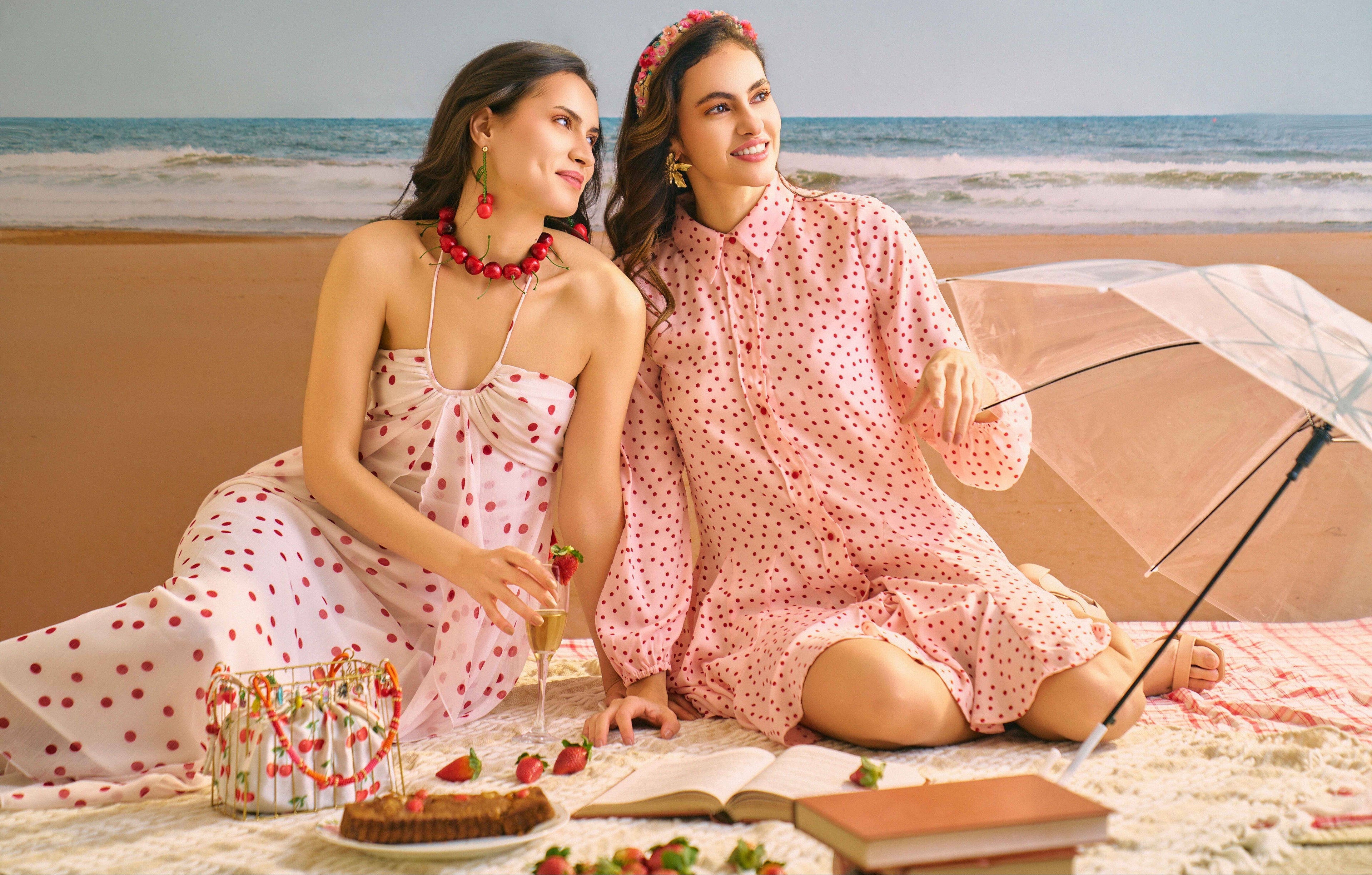 Two women in matching pink polka dot dresses sitting on a beach with an umbrella and snacks.