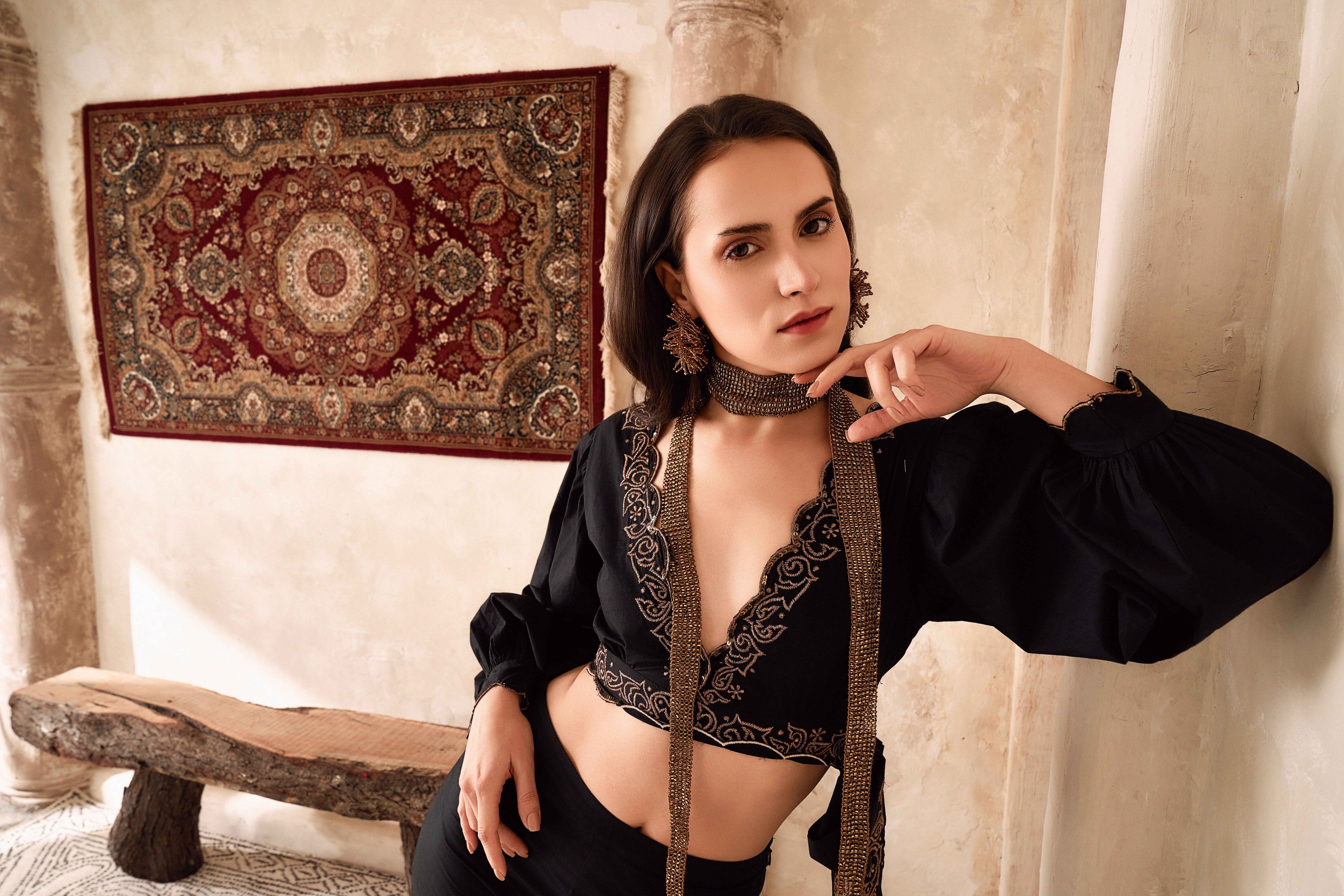 Woman in a black outfit with intricate designs in a room with a patterned rug and wooden bench.